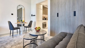 A modern living room with a gray sofa, a dining area with two chairs and a small table, and a kitchen visible through an open doorway. - Chic Studio | Pool View Balcony | Avelon Arjan (Dubai)
