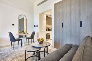 Ein modernes Wohnzimmer mit einem grauen Sofa, einem Essbereich mit zwei Stühlen und einem kleinen Tisch sowie eine durch eine offene Tür einsehbarer Küchenbereich.