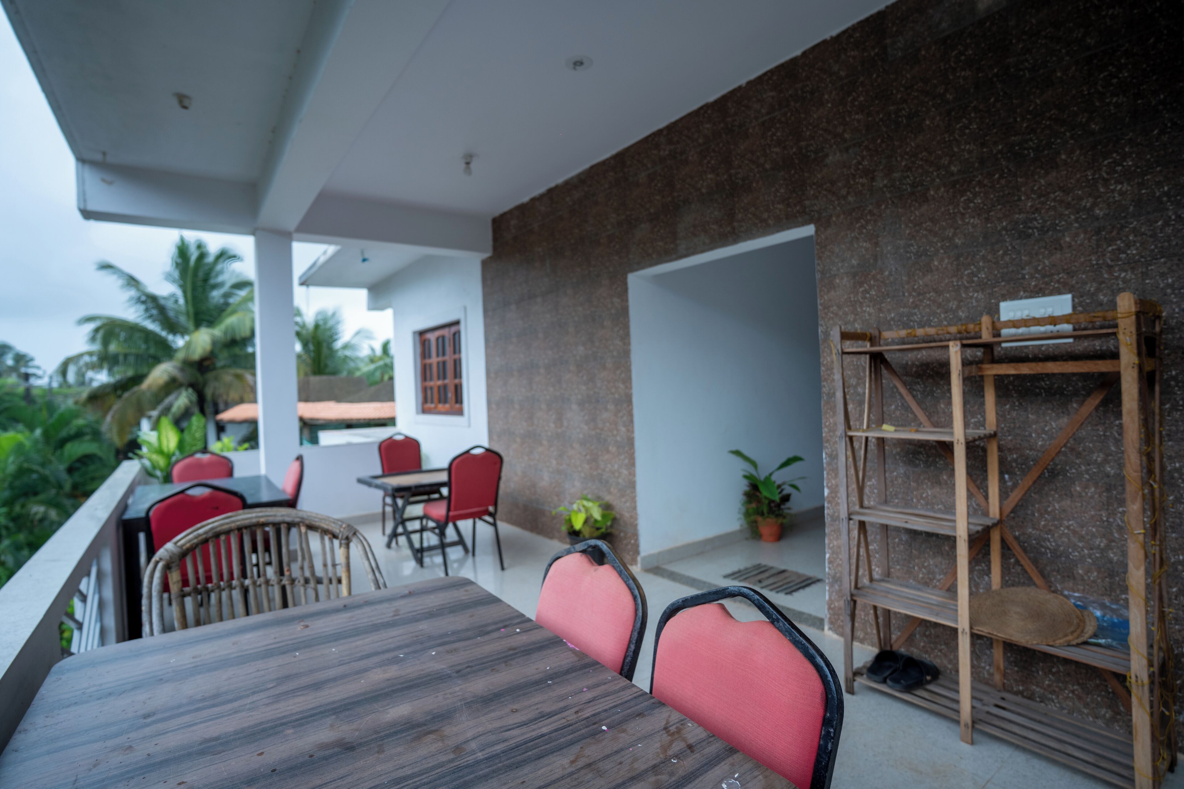 A covered outdoor seating area with a wooden table and red chairs, a wooden shelf unit, and a view of a building with a tiled roof.