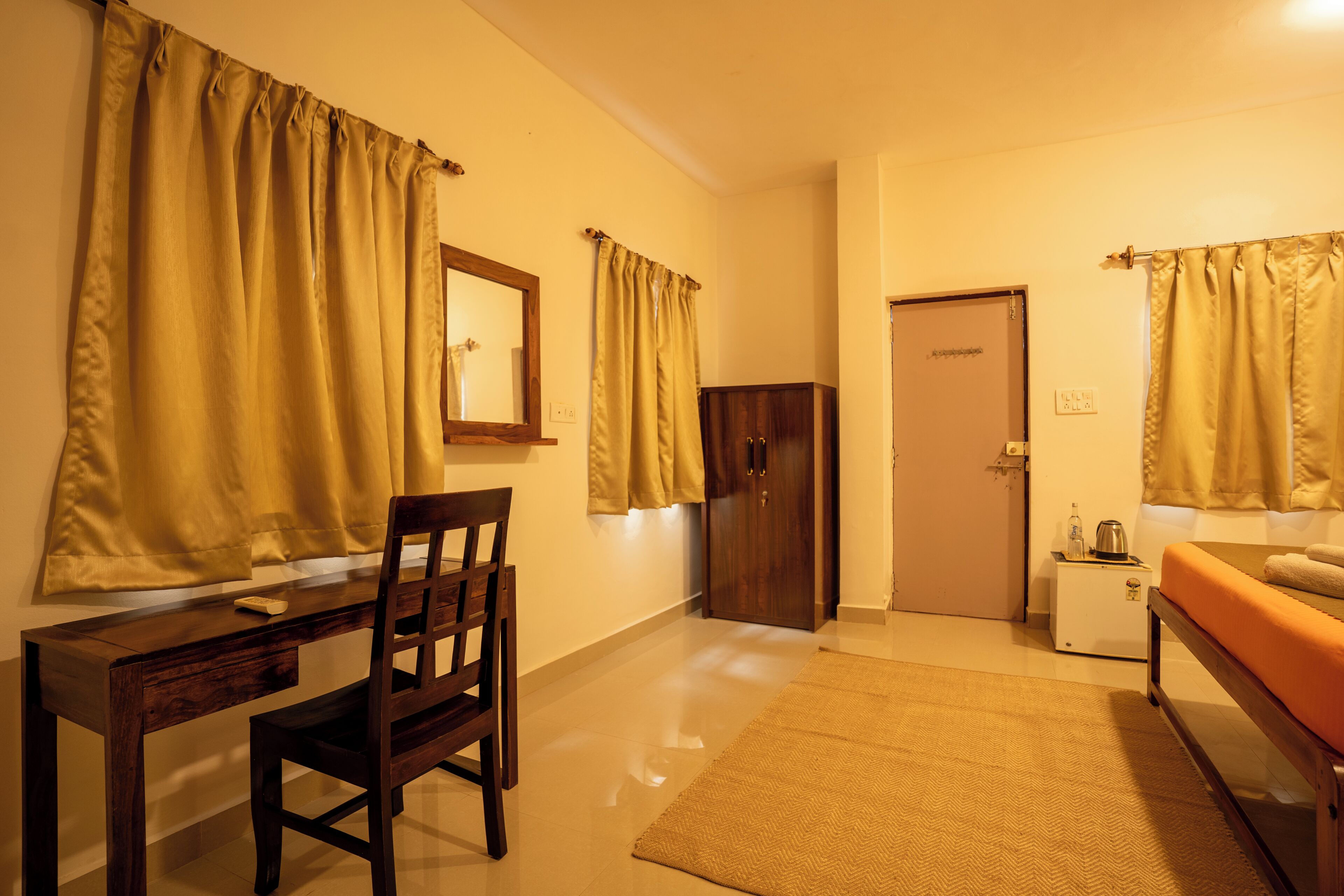 A room with a wooden desk, chair, and a bed with an orange bedspread. There is a wardrobe, a small table with a kettle, and a door in the background.