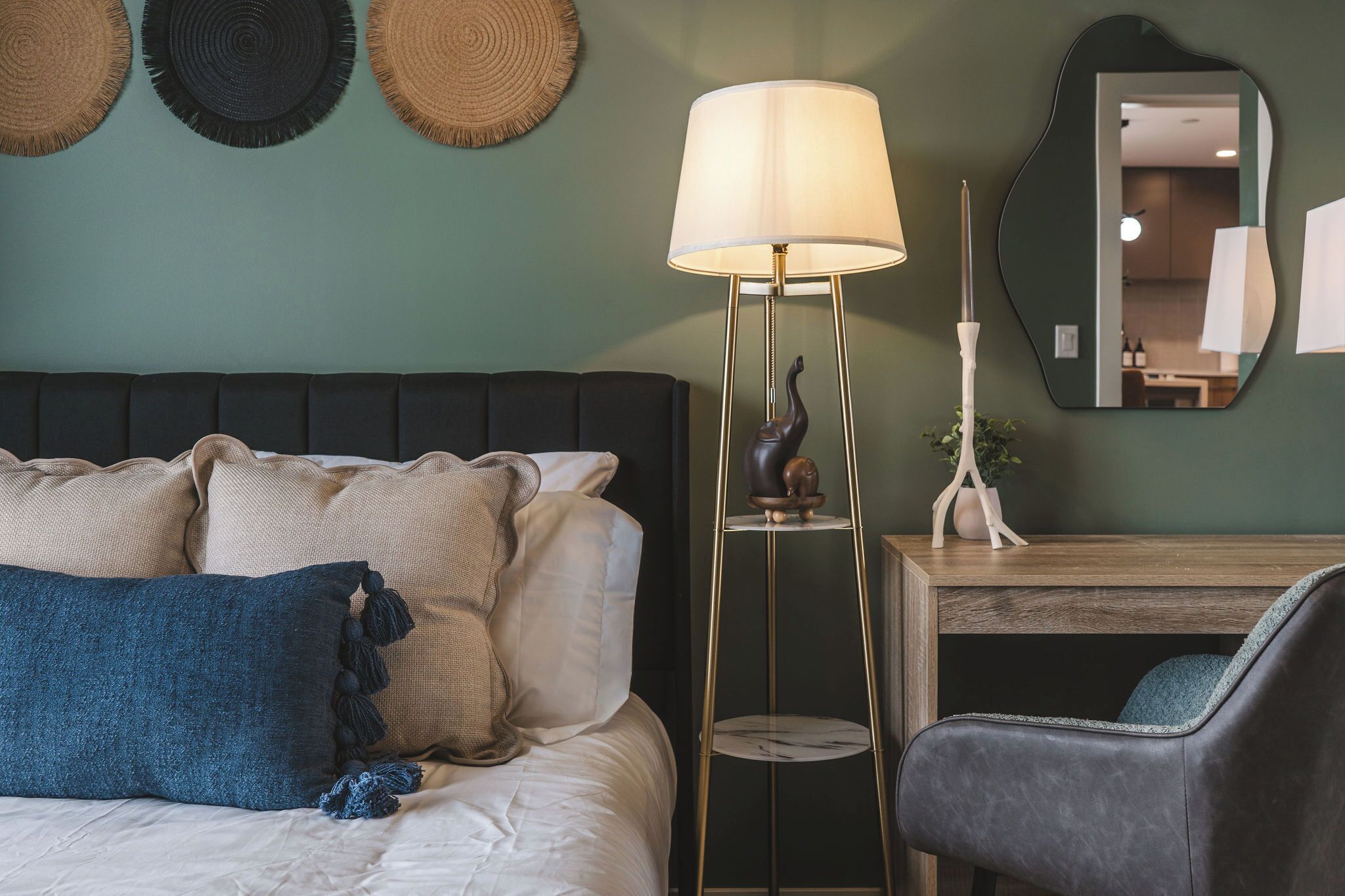 A bedroom with a bed, pillows, a floor lamp, a chair, a mirror, and a small table.
