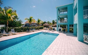 Ein Swimmingpool mit Liegestühlen, ein blaues Gebäude mit Balkonen und Palmen.