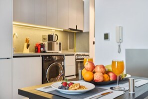 Une cuisine moderne équipée d’un lave-linge, d’un four à micro-ondes et d’une machine à café. On y trouve un panier de fruits contenant des pommes et des oranges, ainsi qu’un verre de jus d’orange sur le comptoir.