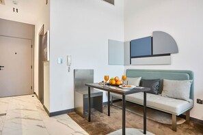 Ein modernes Hotelzimmer mit einer Couch, einem kleinen Tisch mit Obst und Getränken, einem wandmontierten Spiegel und einer Tür.