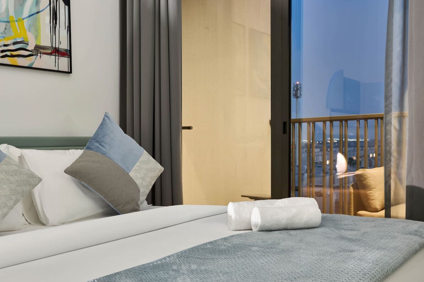 Ein Hotelzimmer mit Bett, Kissen, Decke, Blick auf die Stadt durch eine Balkontür und einem Gemälde an der Wand.
