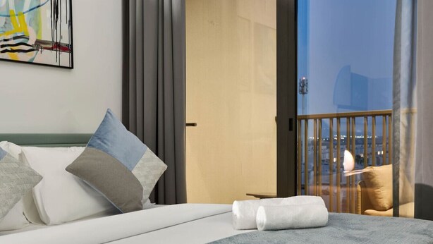 A hotel room with a bed, pillows, a blanket, a view of the city through a balcony door, and a painting on the wall. - Stylish Modern Studio | MAG 910, Meydan Community (Dubai)