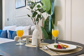 A breakfast setting with a table, two glasses of orange juice, a plate with a strawberry and a slice of cake, and a vase with green plants. - Chic Studio w/ Canal View – Business Bay (Dubai)