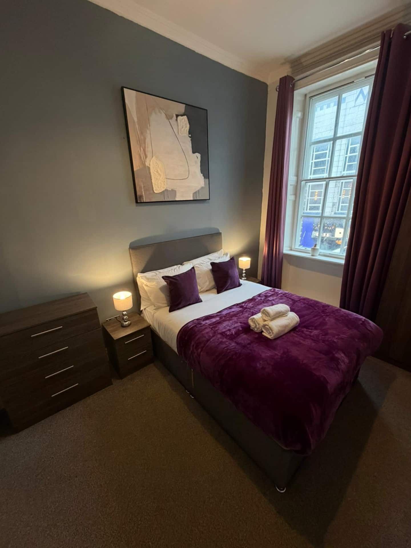 A hotel room with a bed, a nightstand, a drawer unit, a window with curtains, and a painting on the wall.