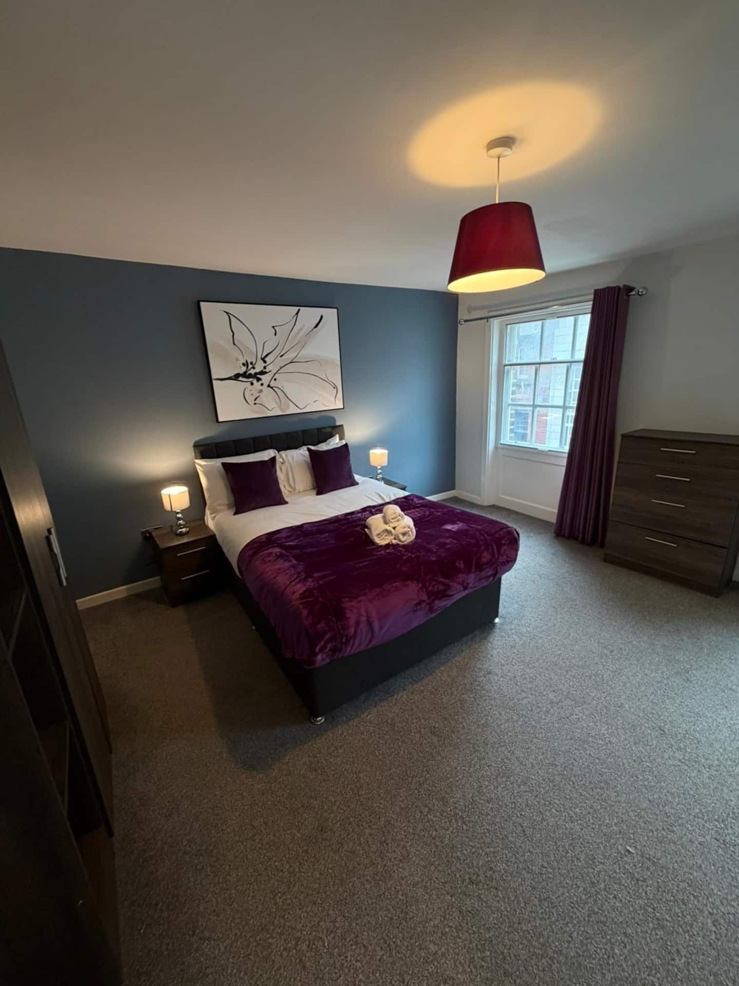 A bedroom with a bed, bedside lamps, a window with curtains, a dresser, and a ceiling light fixture.