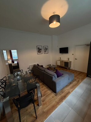 Una sala moderna con un sofá modular, una mesa de comedor con sillas, un televisor de pantalla plana montado en la pared y una lámpara colgante.