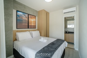Quarto de hotel moderno com cama bem arrumada, pintura de um pôr do sol e vista para a área da cozinha.