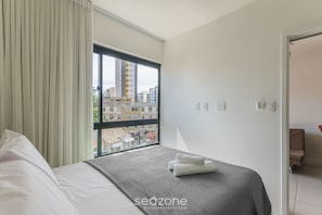 Una habitación de hotel con una cama, un ventanal con cortinas, vistas a edificios y una puerta que da a otra habitación.