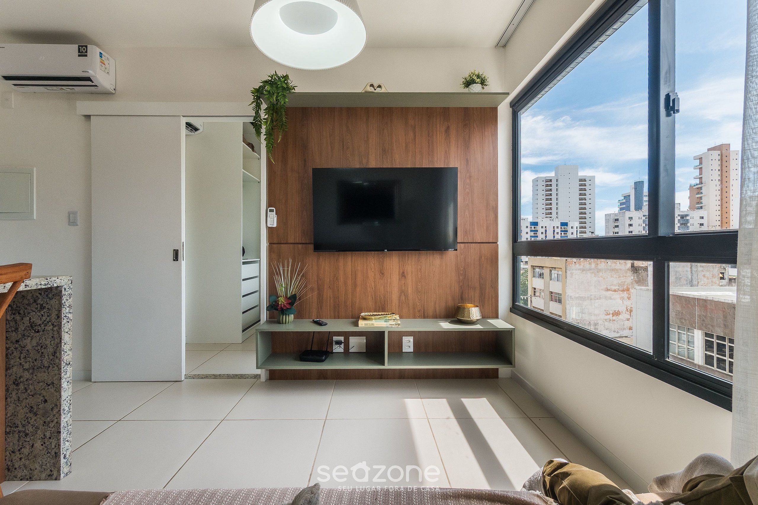 A modern living room with a large TV mounted on a wooden wall, a glass coffee table, and a view of the city through a large window.