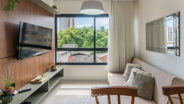 Un soggiorno moderno con una TV a parete, un divano con cuscini, una panca decorata con oggetti e un'ampia finestra che si affaccia su vegetazione e palazzi.