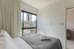 Una habitación de hotel con una cama, un ventanal con cortinas, vistas a edificios y una puerta que da a otra habitación.