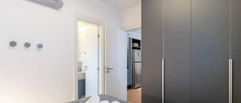 A modern bedroom with a large bed, a wardrobe, and a kitchenette in the background.