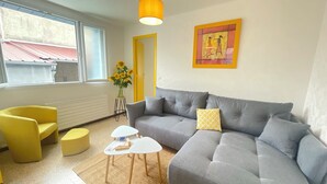 A modern living room with a grey sofa, a yellow armchair, and a small white table. There is a vase with sunflowers on a side table, and a painting on the wall.