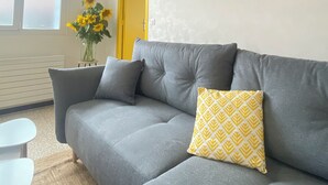 A grey sofa with a patterned pillow, a yellow-framed painting on the wall, and a vase with yellow flowers on a small table.