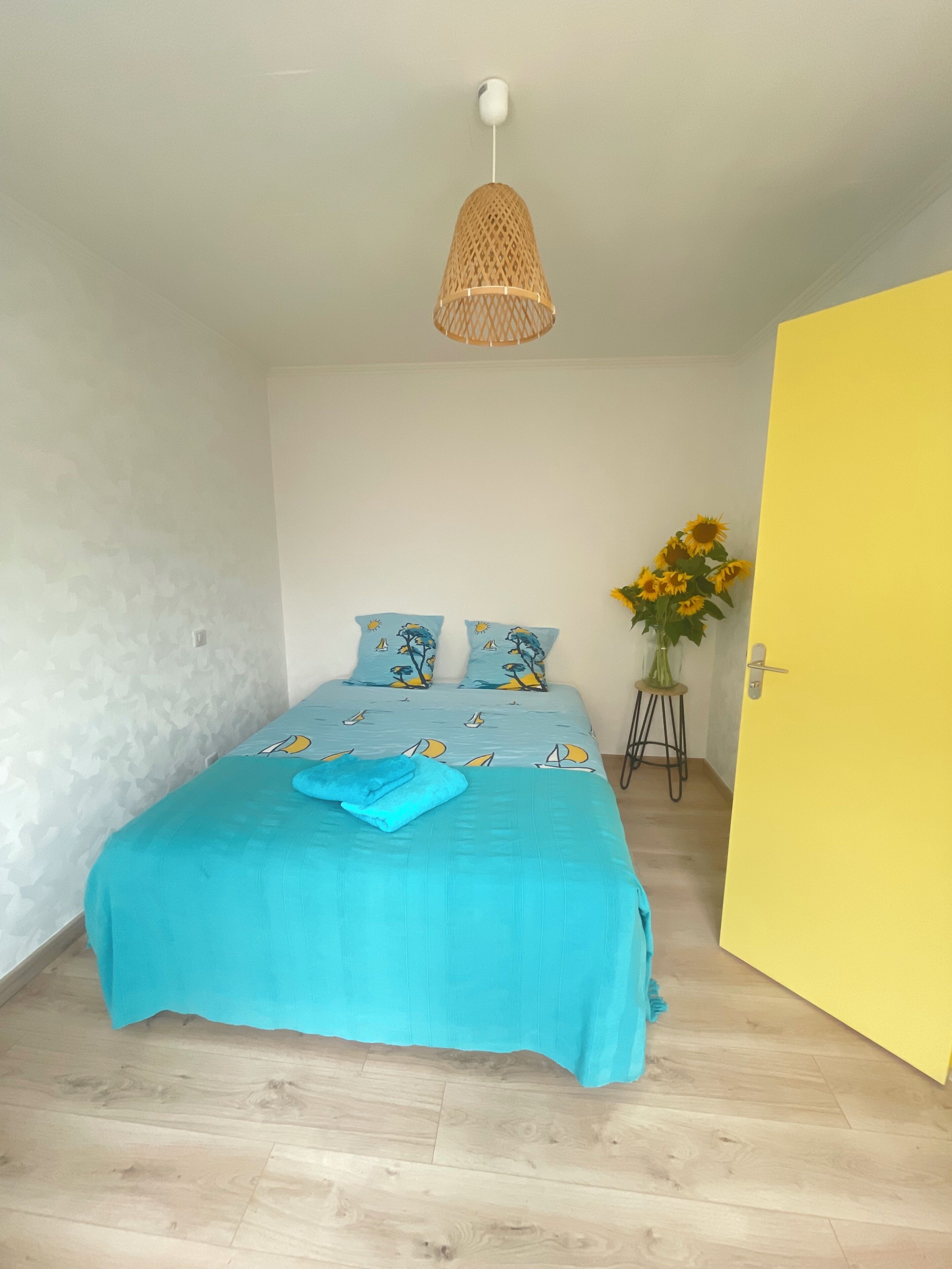 A bedroom with a bed, a hanging light fixture, a door, and a vase with sunflowers.