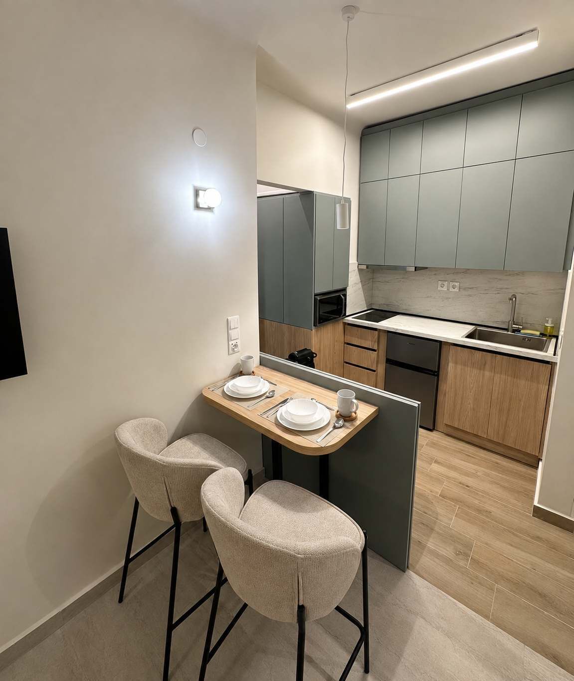 Eine moderne Küche mit grauen Schränken, einer Holz Arbeitsplatte und einem eingebauten Backofen. An einem kleinen Esstisch stehen zwei Stühle, Teller und Besteck.