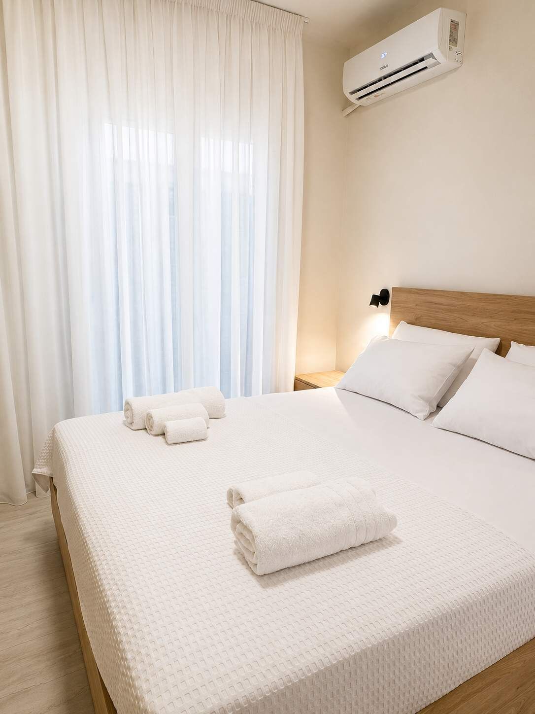 Ein Hotelzimmer mit einem Bett, weißen Leinen, einem Holz-Kopfschild, einem Nachttisch, einer Wand-Klimaanlage und durchsichtigen Vorhängen.