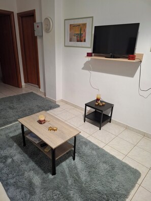 Ein Wohnzimmer mit Couchtisch aus Holz, einem kleinen schwarzen Beistelltisch mit Kerze, einem Fernseher auf einer Holzablage und einem flauschigen grauen Teppich.