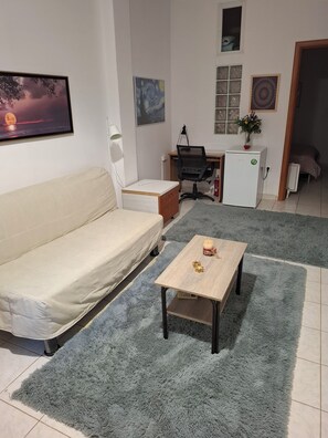 Ein Wohnzimmer mit einer Couch, einem Couchtisch, einem Schreibtisch mit Stuhl, einem kleinen Kühlschrank und einem Fenster mit Jalousien.