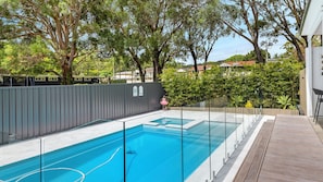 A rectangular swimming pool with a glass barrier, surrounded by a wooden deck and a metal fence. - Sandy Toes – Three Lux Retreats with Heated Pool (Nelson Bay)