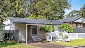 A residential property with a covered porch, a stone facade, and a fenced yard with various plants. - Sandy Toes – Three Lux Retreats with Heated Pool (Nelson Bay)
