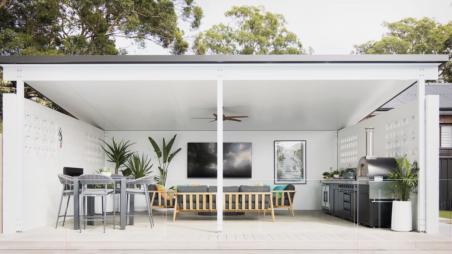 A covered outdoor area with a dining table, chairs, a ceiling fan, and a kitchen area with a grill.