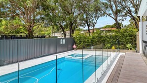 A rectangular swimming pool with a glass barrier, surrounded by a wooden deck and a metal fence. - Sandy Toes – Lux Retreat with Heated Pool (Nelson Bay)