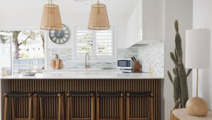 A modern kitchen with a central island, two hanging pendant lights, a clock, and a view of the outdoors. - Sandy Toes – Lux Retreat with Heated Pool (Nelson Bay)