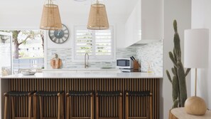 A modern kitchen with a central island, two hanging pendant lights, a clock, and a view of the outdoors. - Sandy Toes – Lux Retreat with Heated Pool (Nelson Bay)