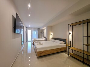 A modern hotel room with a large bed, a TV mounted on the wall, and a wooden shelf unit. - Odyssey Superior Junior Suite (Paralia)