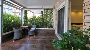 Ein überdachter Patio-Bereich mit Tisch und zwei Korbstühlen, einer Ziegelwand und einer großen Schiebeglasdoor, die zu einem Schlafzimmer führt.