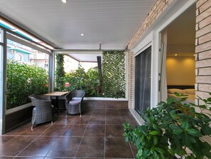 A covered patio area with a table and two wicker chairs, a brick wall, and a large sliding glass door leading to a bedroom. - Odyssey Superior Junior Suite (Paralia)