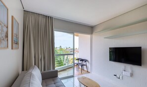 Uma sala de estar moderna com sofá, televisão e varanda com vista para áreas verdes e edifícios.