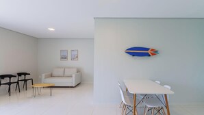 Un salon moderne avec un canapé blanc, deux fauteuils noirs, une petite table ronde et une table à manger avec quatre chaises. Deux tableaux encadrés sont accrochés au mur.