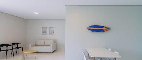 A modern living room with a white sofa, two black chairs, a small round table, and a dining table with four chairs. There are two framed pictures on the wall.