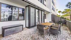 A patio with a wooden table and wicker chairs, a bench, and a view of a building with large windows and a balcony. - 2BD Condo with Pool and Patio Ground Floor (Siesta Key)