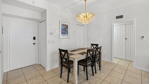 A dining area with a wooden table and chairs, a chandelier, and a door with a security camera. - 2BD Condo with Pool and Patio Ground Floor (Siesta Key)