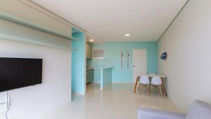 Uma sala de estar moderna com televisão de tela plana, um espaço de jantar com mesa e cadeiras, e uma cozinha com armários e uma janela.