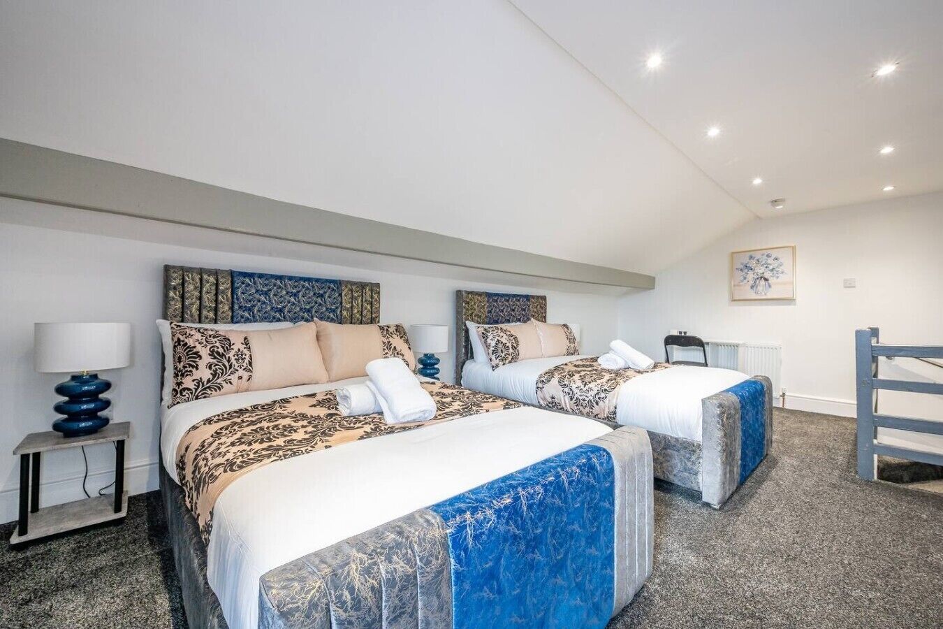 A hotel room with two beds, a small table, a chair, and a painting on the wall.