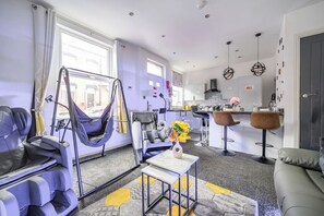 A modern living room with a sofa, a hanging chair, a coffee table, and a kitchen area with bar stools. - Stylish 2-Bedroom House in Leeds (West Yorkshire)