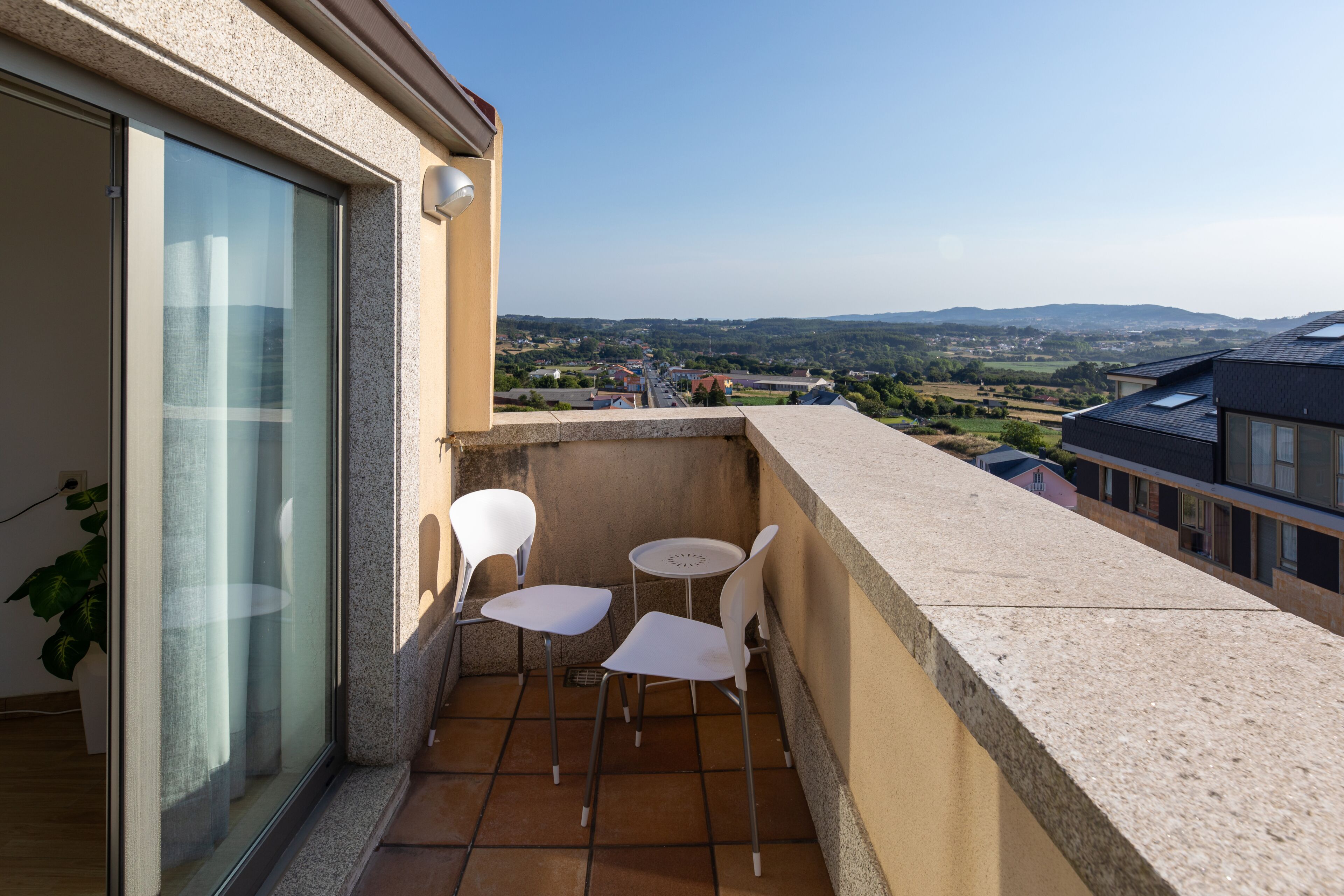 Un balcon avec deux chaises blanches et une petite table, d’où l’on peut admirer un paysage pittoresque composé d’une ville et de collines.