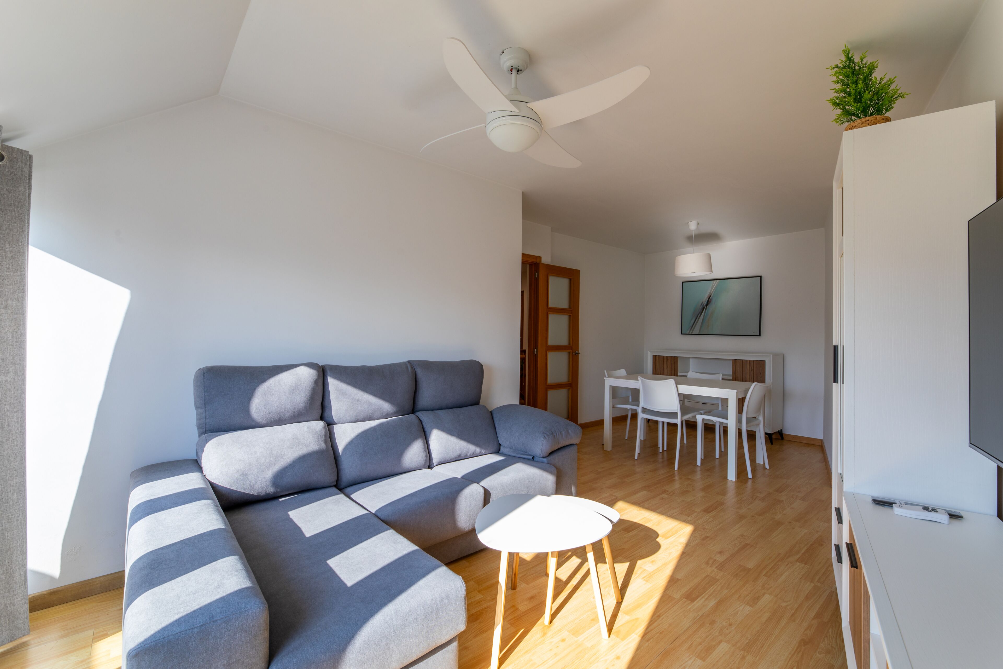 A modern living room with a sectional sofa, a small round table, a ceiling fan, a dining area with a white table and chairs, and a TV on a cabinet.