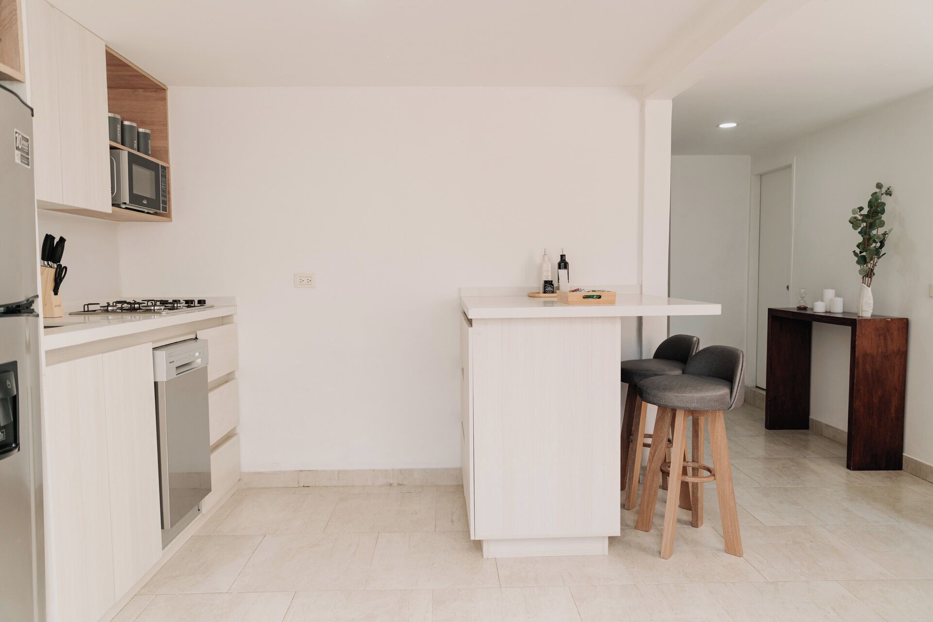 Een moderne keuken met een ingebouwde kookplaat, magnetron en vaatwasser. Er is een witte bartafel met krukken en een kleine houten tafel met flessen en een plant.