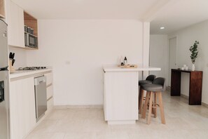 A modern kitchen with a built-in stove, microwave, and dishwasher. There is a white bar counter with stools and a small wooden table with bottles and a plant. - House G with Garage, 15 minutes from JMC Airport—all the comforts in one place. (Rionegro)