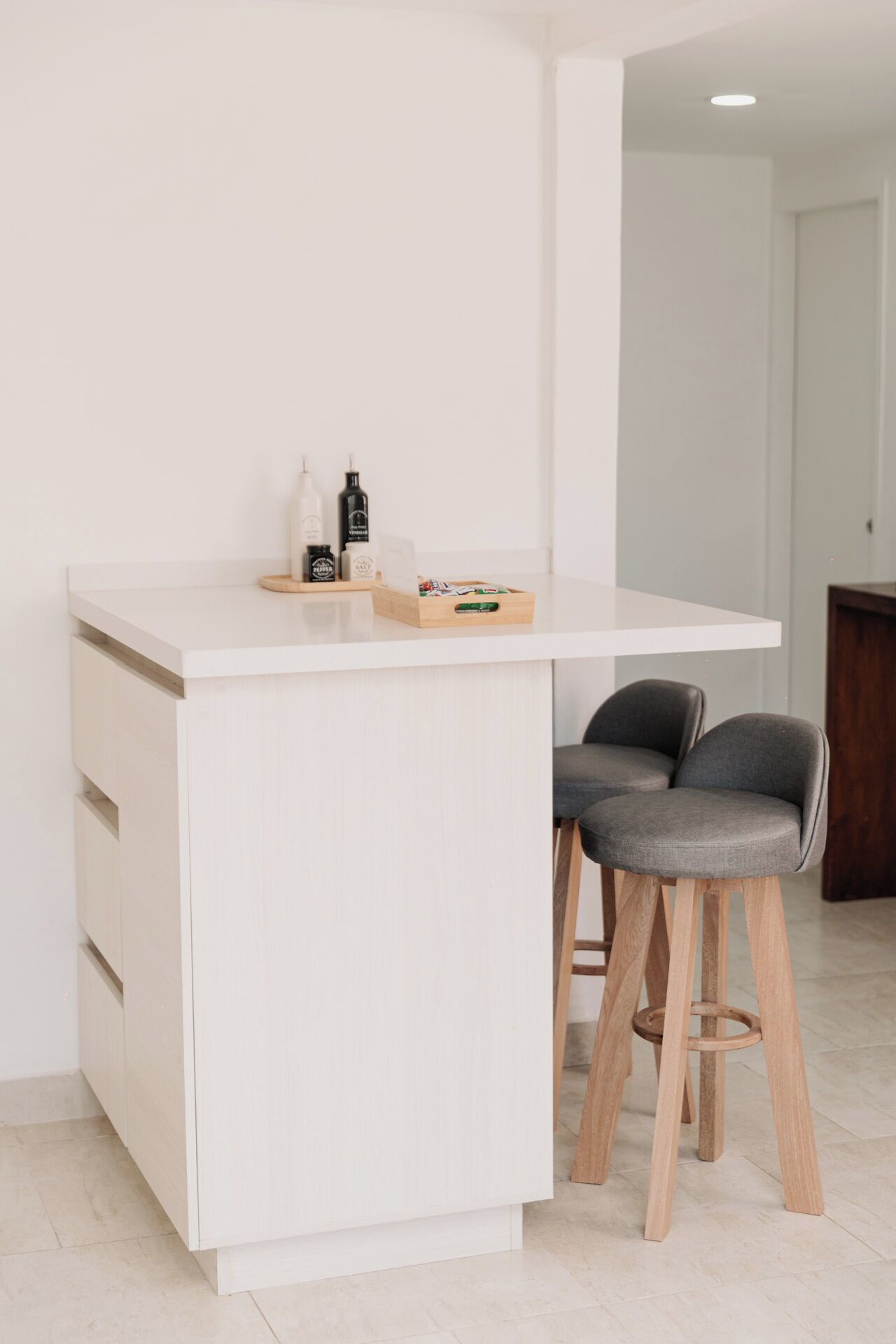Een minimalistische keuken met een wit aanrecht, twee barkrukken en een houten kast.