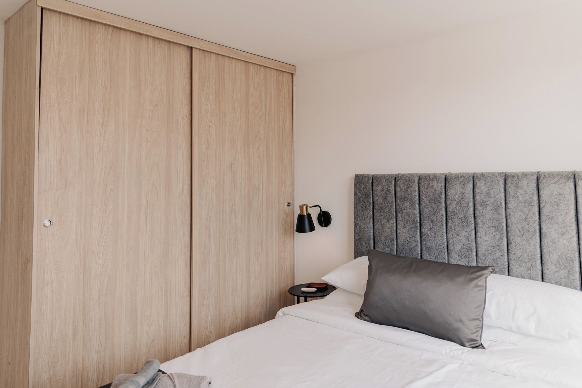 Ein Schlafzimmer mit großem Kleiderschrank, einem Bett mit weißer Bettwäsche, einem grauen Kopfteil und einem kleinen Nachttisch mit Lampe.